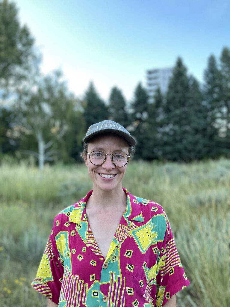 Cass pictured with a field behind them, wearing a colorful shirt, baseball cap, and glasses. 